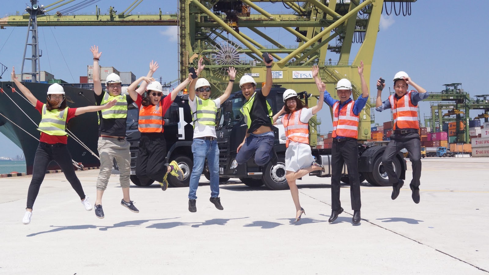 Team in hard hats and hi-vis vests jumping in front of a prime mover truck at a container terminal