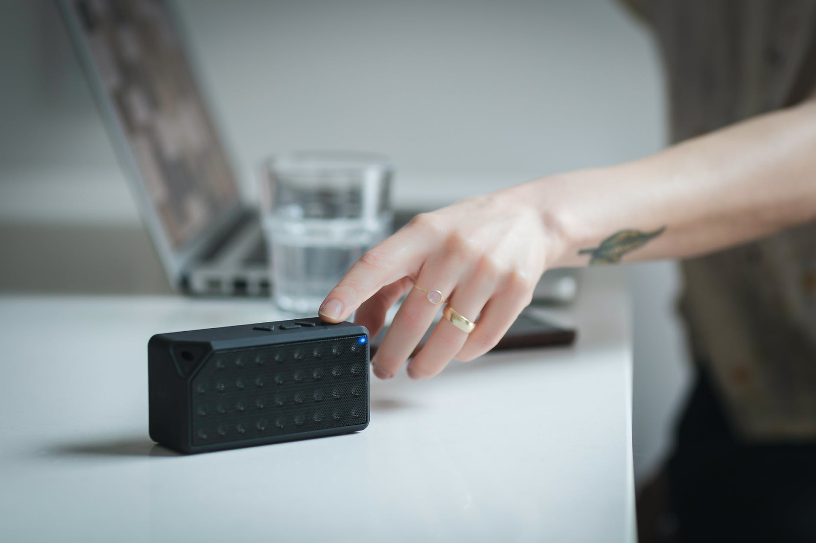A hand pressing the power button on a small portable Bluetooth speaker on a desk