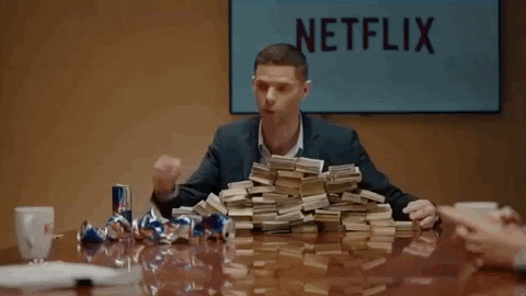 A man in a boardroom thumps the table beside towering stacks of banknotes
