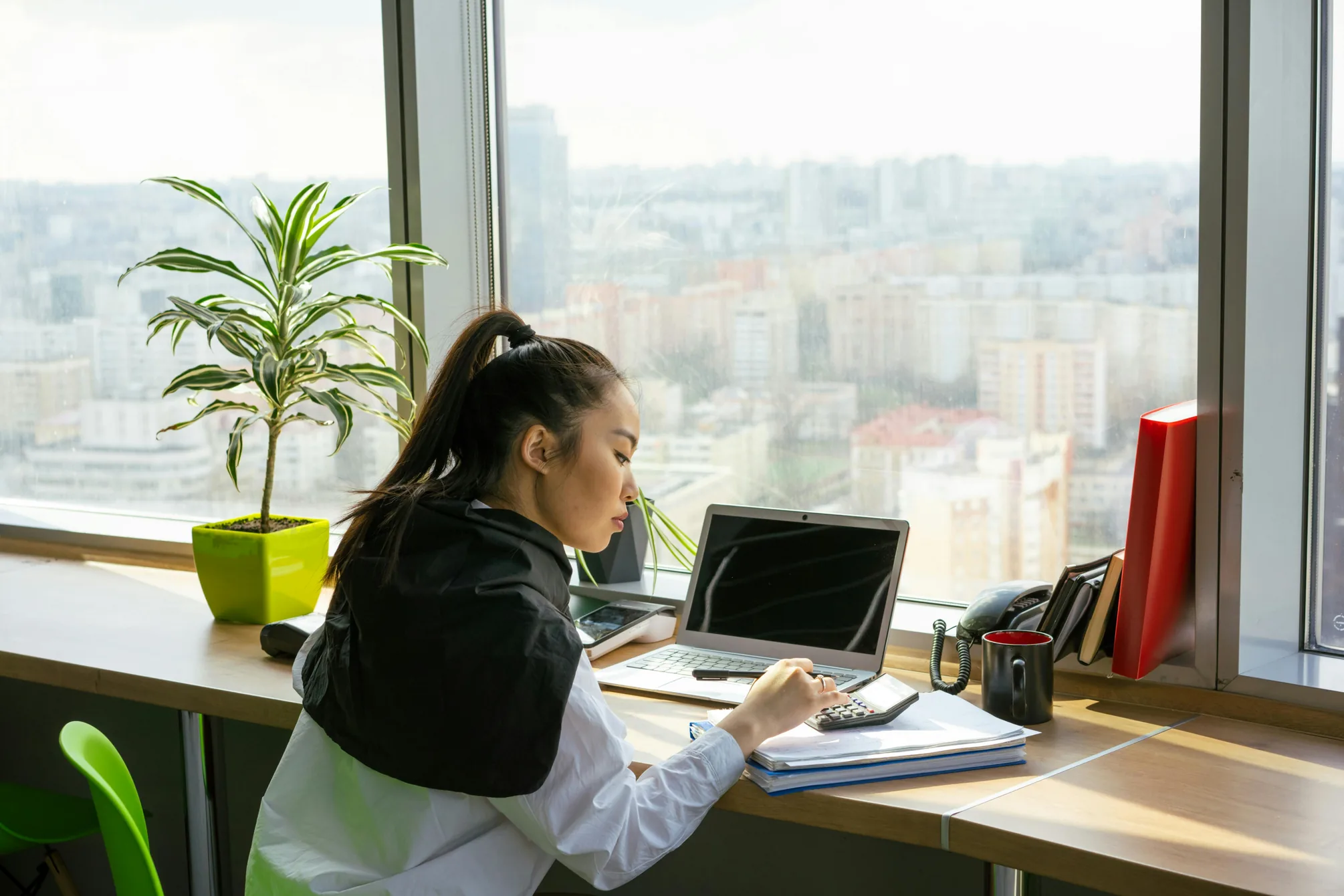 A person working at a laptop at a desk by a window, with a calculator and papers beside them